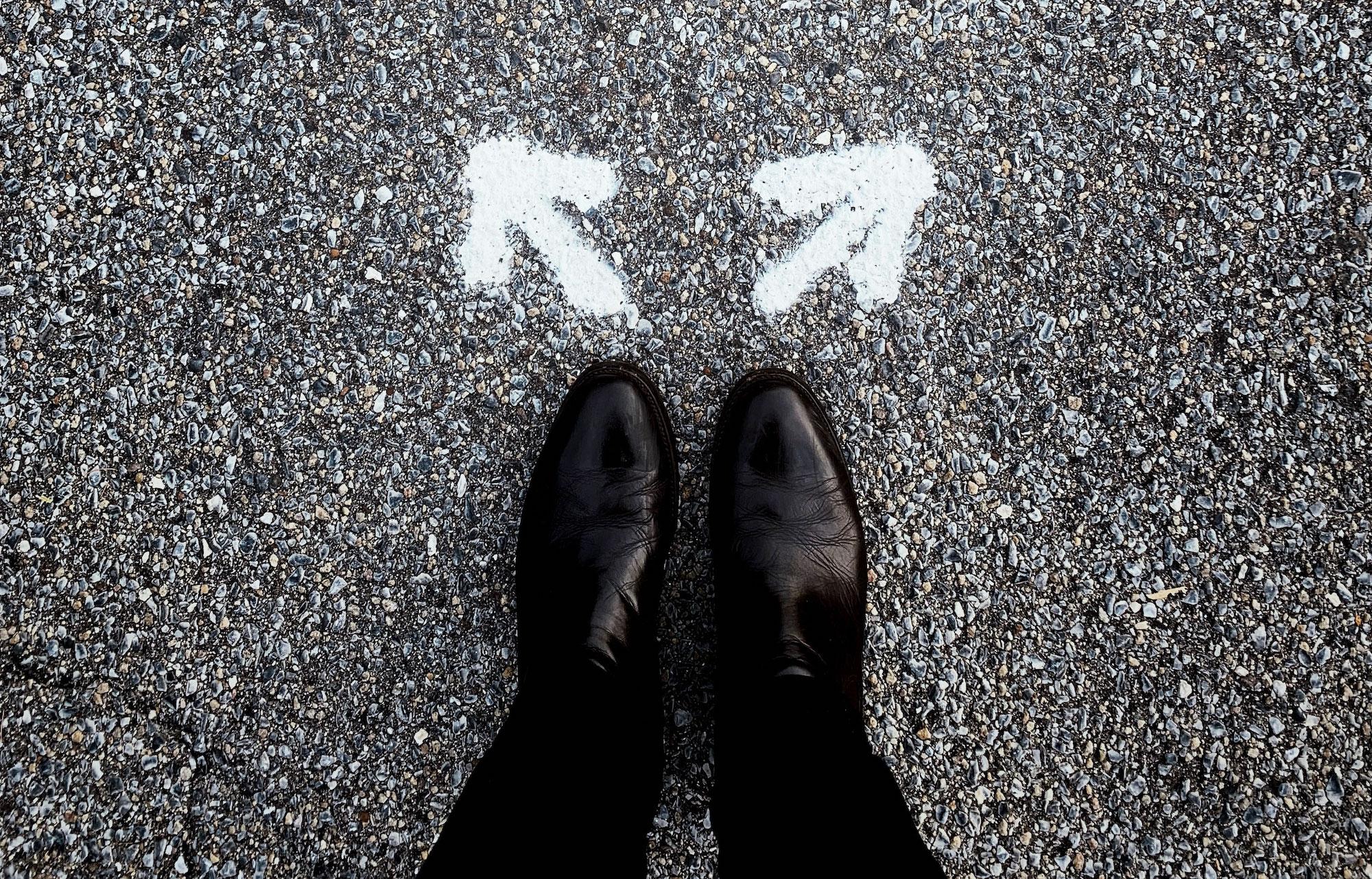 Two feet wearing black boots stand on concrete. In front of the toes, white arrows point in opposite directions.