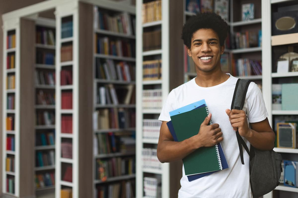 A future Houston teacher standing in his campus library. He’s wearing a white T-shirt and holding books in one hand and a backpack on his shoulder with the other. 